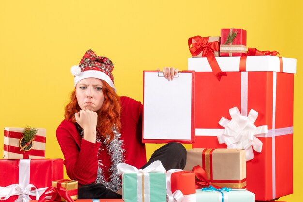 Vista frontal de la mujer joven sentada alrededor de diferentes regalos de Navidad en la pared amarilla