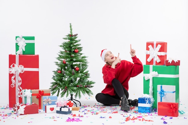 Vista frontal de la mujer joven sentada alrededor de diferentes regalos de vacaciones en la pared blanca