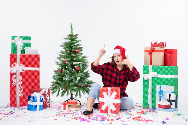 Vista frontal de la mujer joven sentada alrededor de regalos y arbolito de vacaciones en la pared blanca