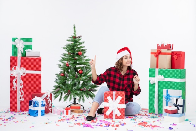 Vista frontal de la mujer joven sentada alrededor de regalos y arbolito de vacaciones en la pared blanca