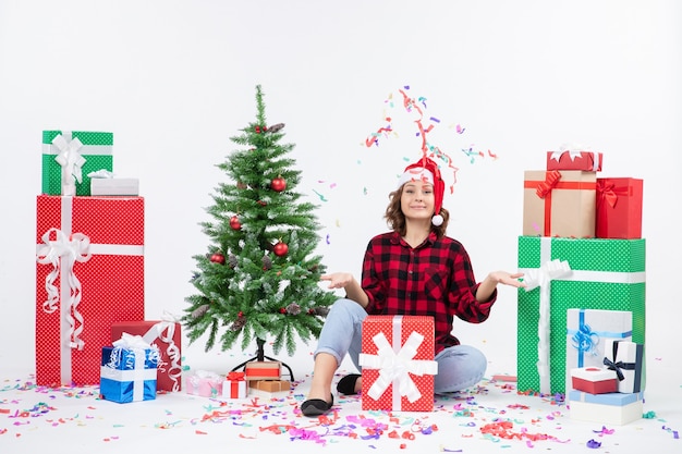 Vista frontal de la mujer joven sentada alrededor de regalos y arbolito de vacaciones en la pared blanca