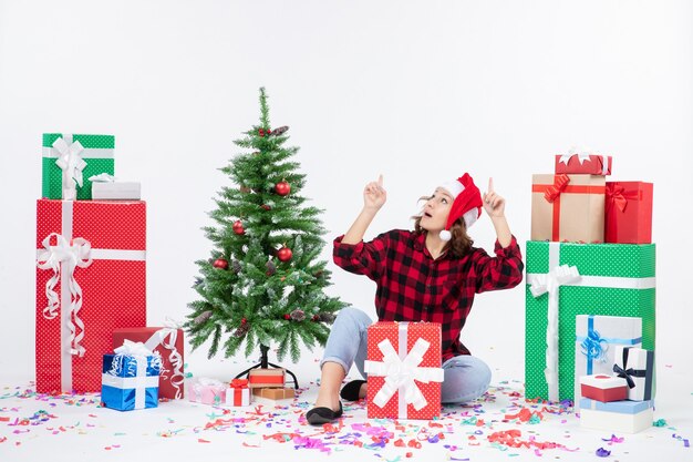 Vista frontal de la mujer joven sentada alrededor de regalos y arbolito de vacaciones en la pared blanca