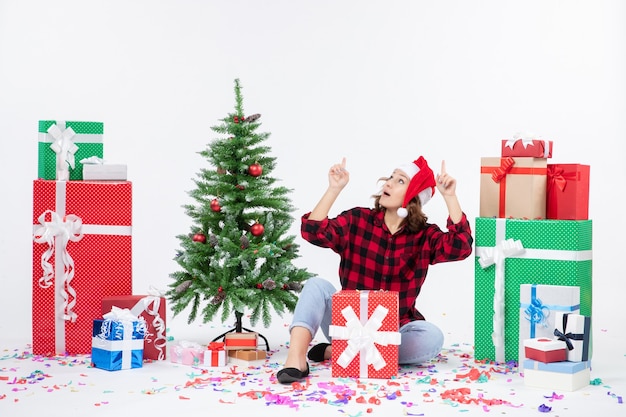 Vista frontal de la mujer joven sentada alrededor de regalos y arbolito de vacaciones en la pared blanca