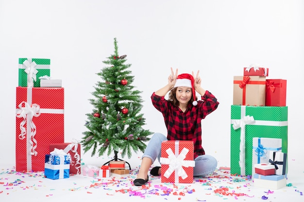 Vista frontal de la mujer joven sentada alrededor de regalos y arbolito de vacaciones en la pared blanca