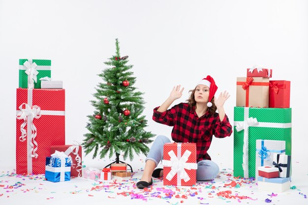 Vista frontal de la mujer joven sentada alrededor de regalos y arbolito de vacaciones en la pared blanca