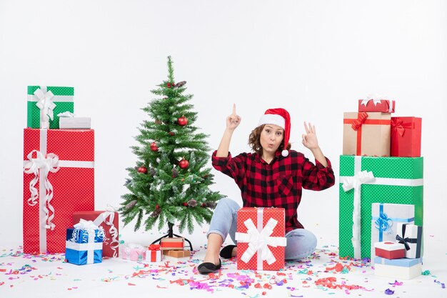 Vista frontal de la mujer joven sentada alrededor de regalos y arbolito de vacaciones en la pared blanca