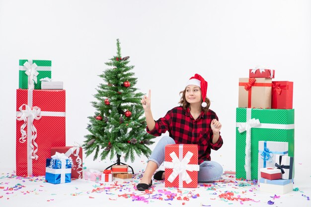 Vista frontal de la mujer joven sentada alrededor de regalos y arbolito de vacaciones en la pared blanca