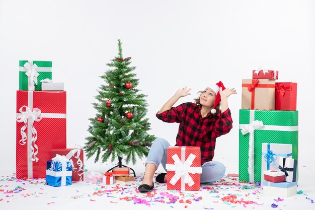 Vista frontal de la mujer joven sentada alrededor de regalos de Navidad y arbolito de vacaciones en la pared blanca