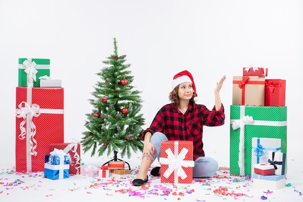 Vista frontal de la mujer joven sentada alrededor de regalos de Navidad y arbolito de vacaciones en la pared blanca