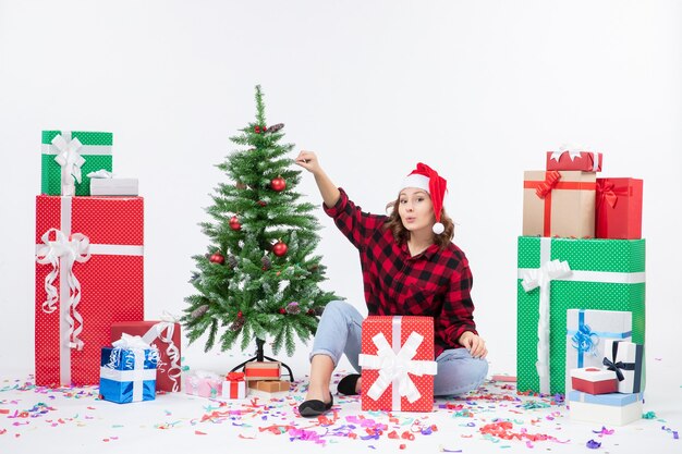 Vista frontal de la mujer joven sentada alrededor de regalos de Navidad y arbolito de vacaciones en la pared blanca