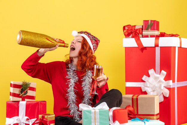 Vista frontal de la mujer joven sentada alrededor de regalos de Navidad y celebrando con champán en la pared amarilla