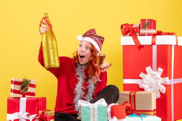 Vista frontal de la mujer joven sentada alrededor de regalos de Navidad y celebrando con champán en la pared amarilla