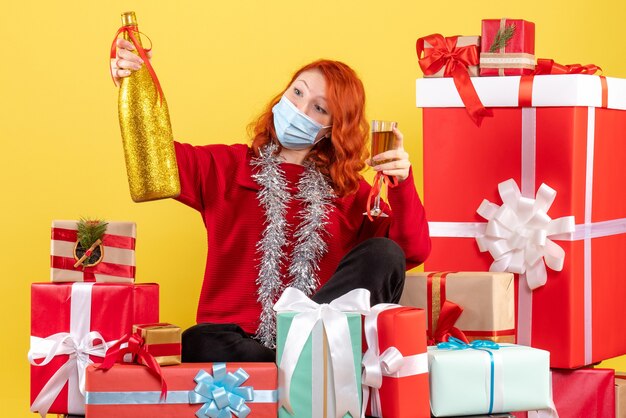 Vista frontal de la mujer joven sentada alrededor de regalos de Navidad en máscara con champán en la pared amarilla