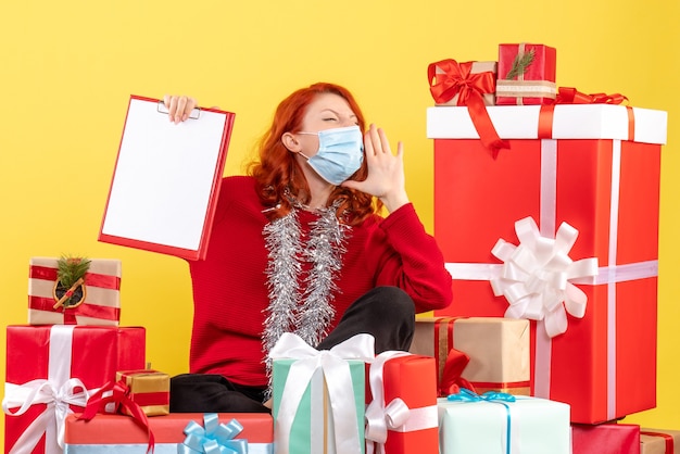 Vista frontal de la mujer joven sentada alrededor de regalos de Navidad en máscara gritando en la pared amarilla