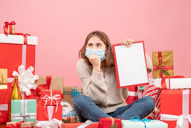 Vista frontal mujer joven sentada alrededor de regalos de Navidad en máscara con nota