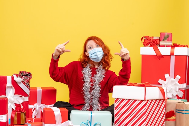 Vista frontal de la mujer joven sentada alrededor de regalos de Navidad en máscara en la pared amarilla