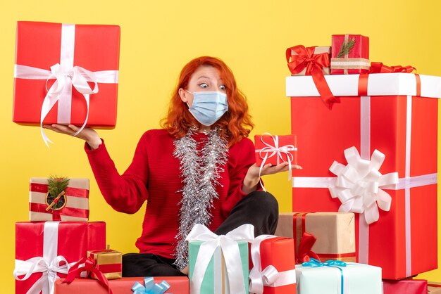 Vista frontal de la mujer joven sentada alrededor de regalos de Navidad en máscara en la pared amarilla