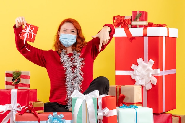 Vista frontal de la mujer joven sentada alrededor de regalos de Navidad en máscara en la pared amarilla
