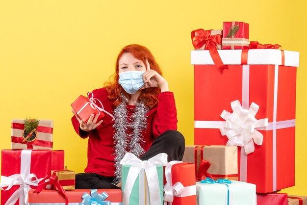 Vista frontal de la mujer joven sentada alrededor de regalos de Navidad en máscara en la pared amarilla