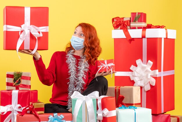 Vista frontal de la mujer joven sentada alrededor de regalos de Navidad en máscara en la pared amarilla