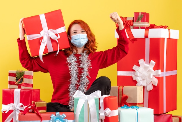 Vista frontal de la mujer joven sentada alrededor de regalos de Navidad en máscara en la pared amarilla