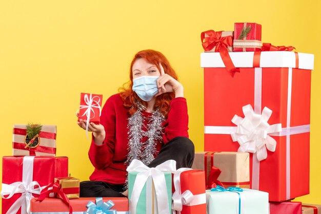 Vista frontal de la mujer joven sentada alrededor de regalos de Navidad en máscara en la pared amarilla