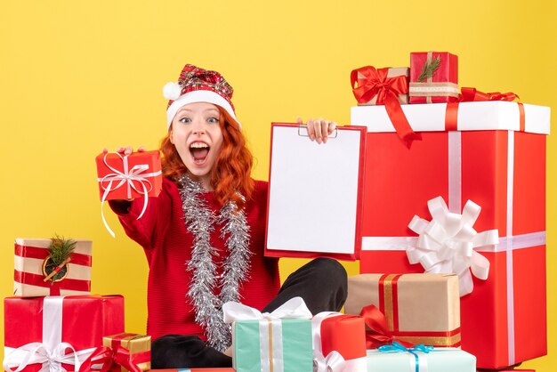 Vista frontal de la mujer joven sentada alrededor de regalos de Navidad con nota de archivo en la pared amarilla