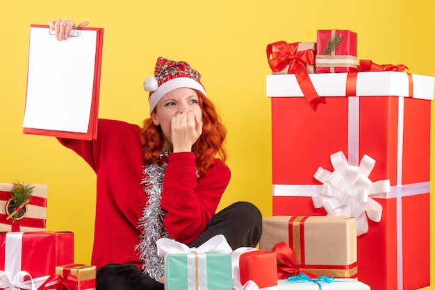 Vista frontal de la mujer joven sentada alrededor de regalos de Navidad con nota de archivo en la pared amarilla