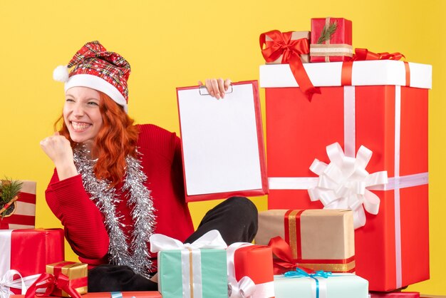 Vista frontal de la mujer joven sentada alrededor de regalos de Navidad con nota de archivo en la pared amarilla