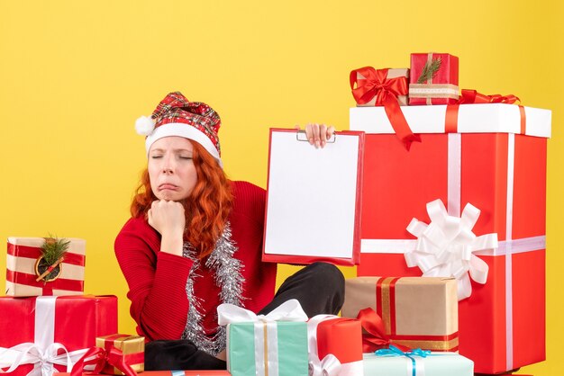 Vista frontal de la mujer joven sentada alrededor de regalos de Navidad con nota de archivo en la pared amarilla