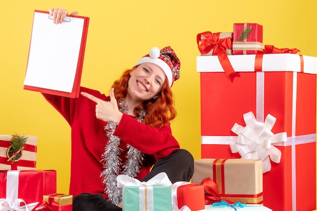 Vista frontal de la mujer joven sentada alrededor de regalos de Navidad con nota de archivo en la pared amarilla