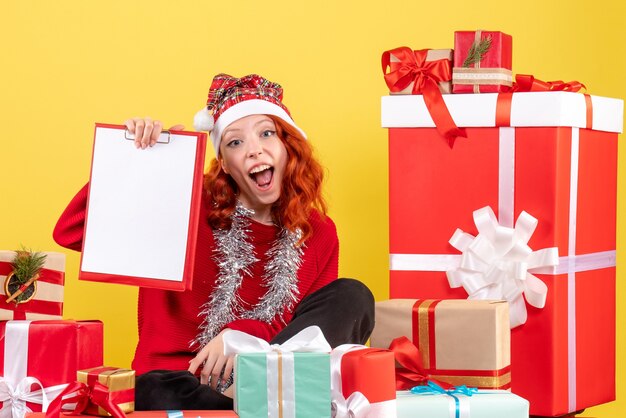 Vista frontal de la mujer joven sentada alrededor de regalos de Navidad con nota de archivo en la pared amarilla