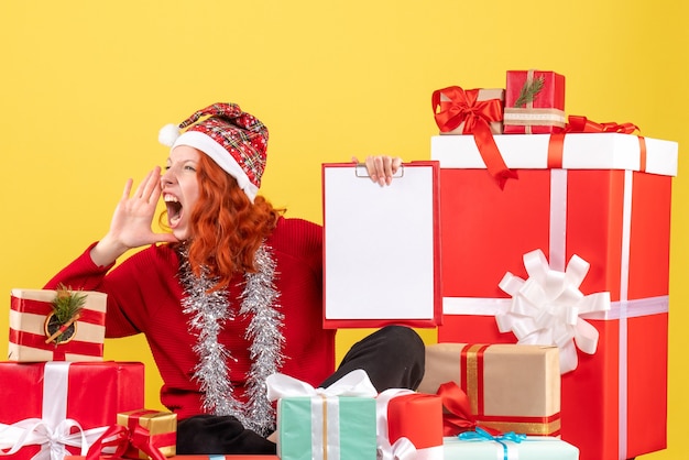 Vista frontal de la mujer joven sentada alrededor de regalos de Navidad con nota de archivo en la pared amarilla