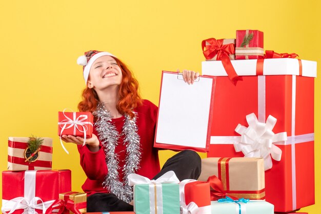 Vista frontal de la mujer joven sentada alrededor de regalos de Navidad con nota de archivo en la pared amarilla