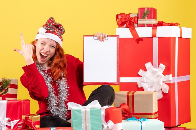 Vista frontal de la mujer joven sentada alrededor de regalos de Navidad con nota de archivo en la pared amarilla