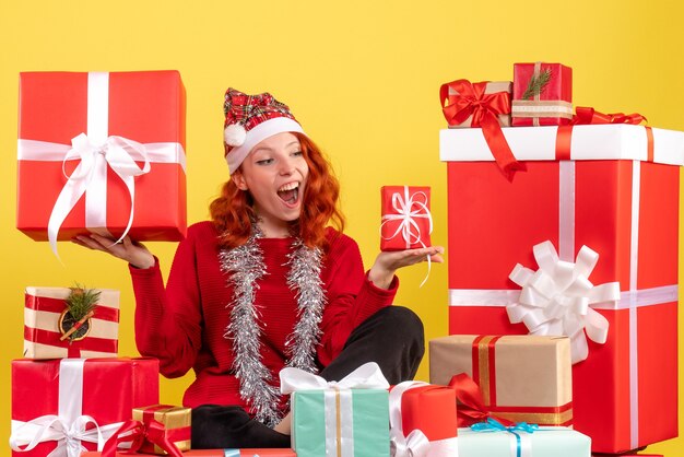 Vista frontal de la mujer joven sentada alrededor de regalos de Navidad en la pared amarilla