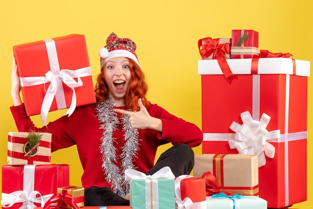Vista frontal de la mujer joven sentada alrededor de regalos de Navidad en la pared amarilla