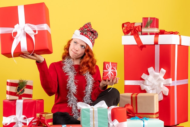 Vista frontal de la mujer joven sentada alrededor de regalos de Navidad en la pared amarilla