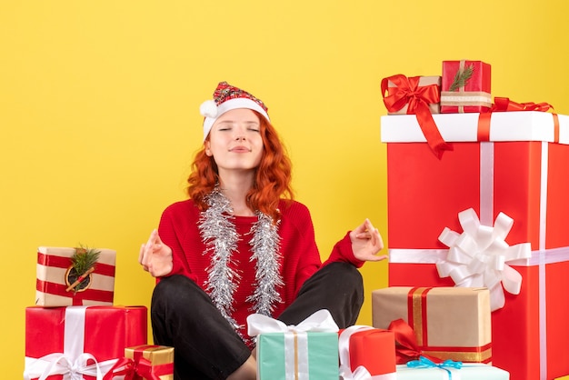 Vista frontal de la mujer joven sentada alrededor de regalos de Navidad en la pared amarilla