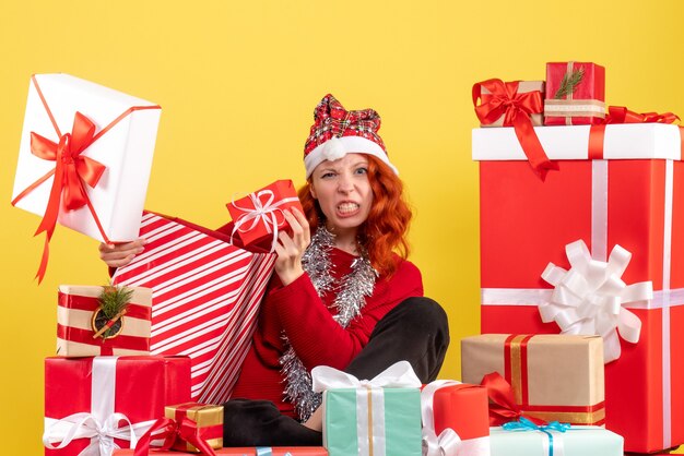 Vista frontal de la mujer joven sentada alrededor de regalos de Navidad en la pared amarilla