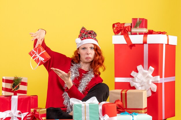 Vista frontal de la mujer joven sentada alrededor de regalos de Navidad en la pared amarilla