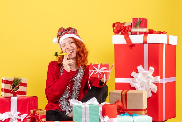 Vista frontal de la mujer joven sentada alrededor de regalos de Navidad en la pared amarilla