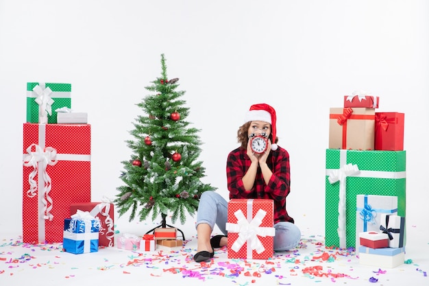 Vista frontal de la mujer joven sentada alrededor de regalos de Navidad sosteniendo relojes en la pared blanca