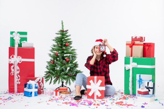 Vista frontal de la mujer joven sentada alrededor de regalos de Navidad sosteniendo relojes en la pared blanca