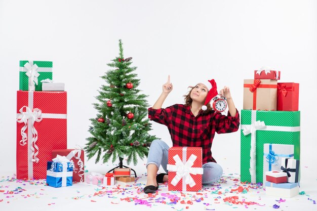 Vista frontal de la mujer joven sentada alrededor de regalos de Navidad sosteniendo relojes en la pared blanca