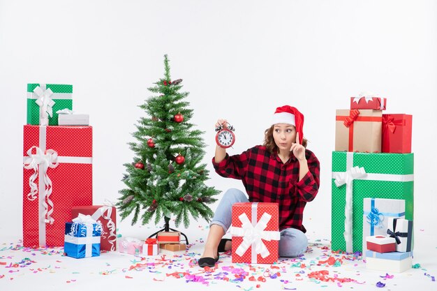 Vista frontal de la mujer joven sentada alrededor de regalos de Navidad sosteniendo relojes en la pared blanca