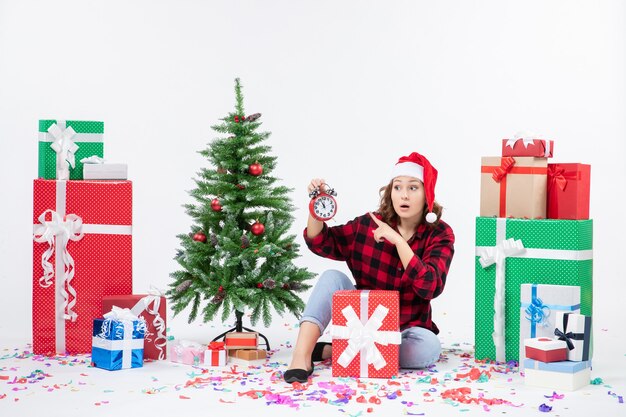 Vista frontal de la mujer joven sentada alrededor de regalos de Navidad sosteniendo relojes en la pared blanca