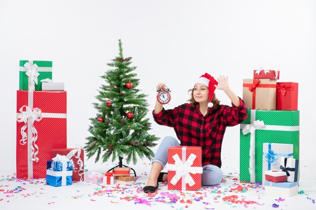 Vista frontal de la mujer joven sentada alrededor de regalos de Navidad sosteniendo relojes en la pared blanca