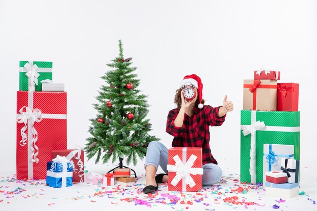 Vista frontal de la mujer joven sentada alrededor de regalos de Navidad sosteniendo relojes en la pared blanca