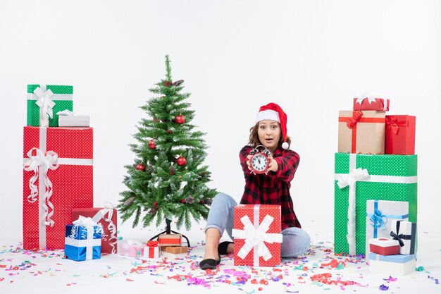 Vista frontal de la mujer joven sentada alrededor de regalos de Navidad sosteniendo relojes en la pared blanca